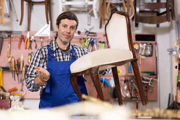 Leisurely middle-aged restorer with hand tool removes old upholstery from vintage chair made of...