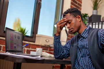 Fototapeta premium African American entrepreneur sits tiredly at desk, holding his head as he analyzes business project plans. Black male manager looking drained after demanding day at work in startup office.
