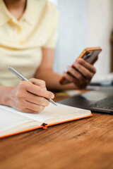 Close-up of a person writing in a notebook while holding a smartphone, with a laptop on a wooden desk.