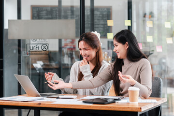 Two young women working together in a modern office, sharing ideas and using a laptop, surrounded by documents and coffee.
