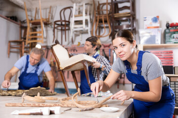 Young woman restorer carefully cleans dirt from carved wooden decor element with hand tool before...