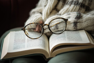 Cozy reading scene with tortoiseshell eyeglasses resting on an open book, held by a person wearing a warm knitted sweater, creating a peaceful and relaxing atmosphere in soft natural light