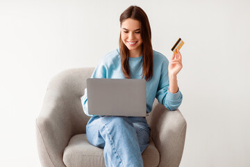Attractive young woman with credit card and laptop sitting in cozy armchair, ordering goods on...