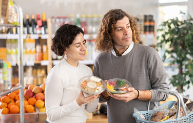 European married couple chooses a packaged poke bowl. Husband discusses the composition of the salad with his wife and looks at the expiration date
