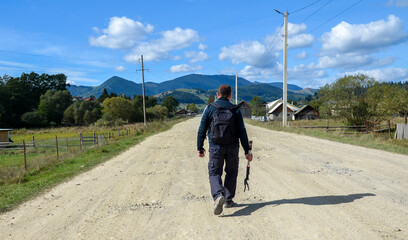Obraz premium Man is walking on a dirt road through a rural countryside setting. Surrounding are green fields, rustic houses, and scenic mountains under a clear blue sky