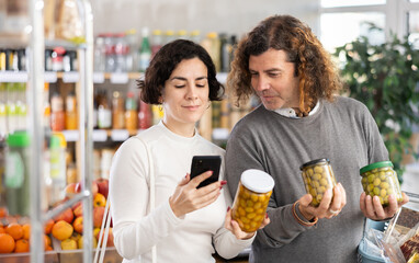 Wife scans the barcode of a jar of olives and shows her husband the product information and expiration date. Couple pays for purchases using a QR code in a supermarket