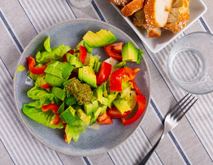 Plate contains portion of salad of fresh seasonal vegetables, complemented by small carafe of olive oil. Salad made from bell pepper, chin cabbage, avocado, pesto dressing