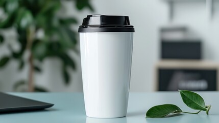 A white travel mug with a black lid sits on a table next to a green plant.