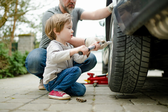 Cute little boy helping his father to change car wheels at their backyard. Father teaching his little son to use tools.