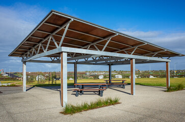 A modern, open-air shelter in a park, with a sloped roof. It houses a picnic table, providing a...