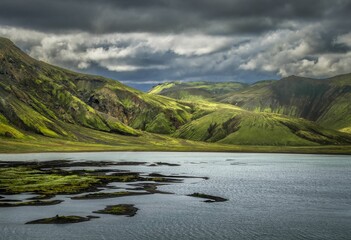Stunning landscape of lush green mountains and a tranquil lake under dramatic clouds.