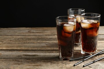 Refreshing cola with ice cubes in glasses and drinking straws on wooden table against black...