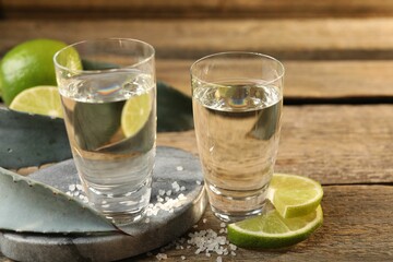 Tequila shots with salt, limes and agave leaves on wooden table, closeup