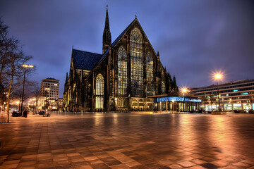 Obraz premium Illuminated Gothic Church Stands Majestically at Night in a City Square with Wet Pavement Reflecting Light