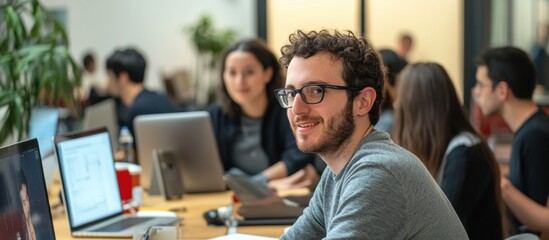 Smiling Man in a Busy Office Environment with Coworkers and Laptop Computers Surrounding Him