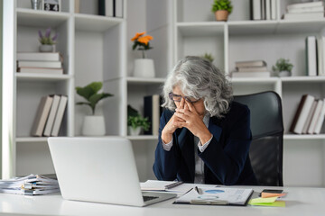 Tired elderly businesswoman with hands on her face while working at her desk in a contemporary office