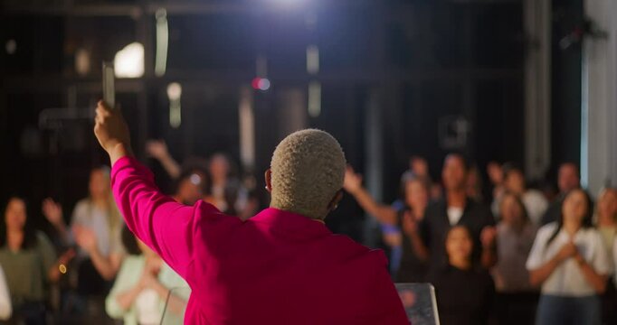 Speaker leading a congregation, back view, holding a Bible with hand raised, dynamic preaching, diverse audience in prayer and worship, indoor church setting, spiritual energy and connection