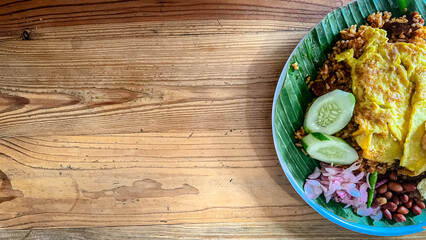 Photo of Aceh Fried Rice made from white rice, eggs, Sumatran spices with pickled onions and fried peanuts, this dish is served on a banana leaf in the shape of a plate with a wooden table in the back