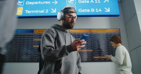 Busy Airport Terminal: Man with Backpack Checks Flight Information on Mobile Phone, Looks at Digital Arrival and Departure Board. Traveler Going on Holiday Trip. Diverse People Walking In Background.