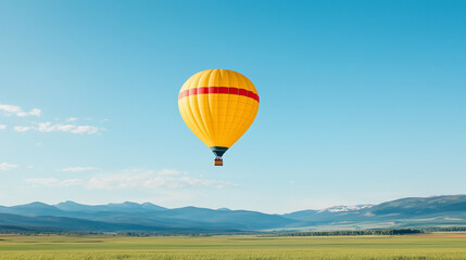 Obraz premium Yellow Hot Air Balloon over Mountains: A vibrant yellow hot air balloon with a red stripe drifts serenely against a clear blue sky, with majestic mountains and a verdant valley stretching out below.