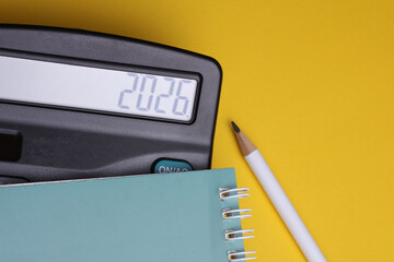Calculator with number 2026 and notebook with pencil on yellow background. Economic analysis,...