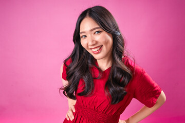 A confident young Asian woman with long black wavy hair wears a red dress and stands with hands on hips against a pink background. She looks directly at the camera with a friendly smile.