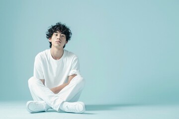 Young Asian Man in White Clothing Sitting Calmly on Light Blue Background