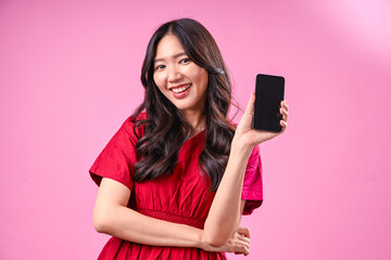 Naklejka premium A cheerful young Asian woman with long black wavy hair wearing a red dress presents a black smartphone with both hands, smiling confidently against a vibrant pink background