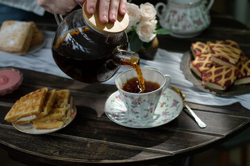 Woman serving tea from teapot in kitchen. Enjoying morning breakfast meal with cup of tea. 