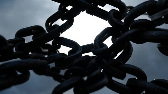 High contrast backlit video, dolly in and out, the camera dollies out from a tight knot of heavy, weathered iron chains against a blinding light source