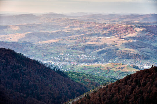 Panoramic photo mountain valley bathed in soft, golden light. Small town nestled in valley, surrounded by rolling hills and fields. Distant mountains fade into horizon, creating sense of depth.