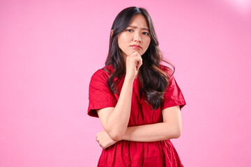 A beautiful young Asian woman with long wavy black hair wears a red dress and looks up thoughtfully while resting her chin on her hand against a pink background, expressing curiosity