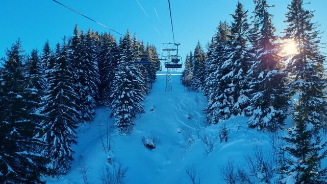 Shot from a ski chairlift ascending a shadowy valley lined with snow-covered fir trees, as sunlight occasionally breaks through on a cold morning at Betelberg in Lenk, Switzerland