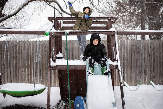 Two kids enjoy snowy fun on a backyard wooden playset and slide