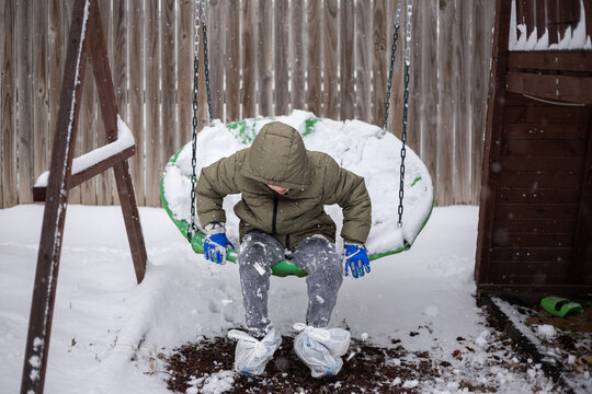 boy  sitting on a snow-covered swing with plastic bags over shoes