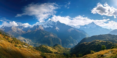 Obraz premium Majestic panoramic view of snow-capped mountains under a vibrant blue sky with wispy clouds and green hills in the foreground showcasing natural beauty.
