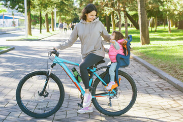 Smiling mother riding a bicycle with her daughter sitting in a child bike seat, enjoying a day...