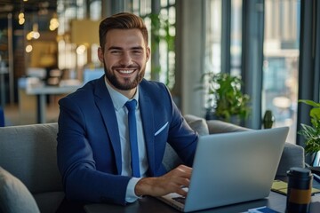 Fototapeta premium A professional image of a young man in business attire, sitting with a laptop on his lap and smiling at the camera. He appears to be working in an office setting.