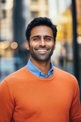 Fototapeta premium An image of a man wearing an orange sweater and blue shirt, smiling brightly at the camera against a blurred city street backdrop.
