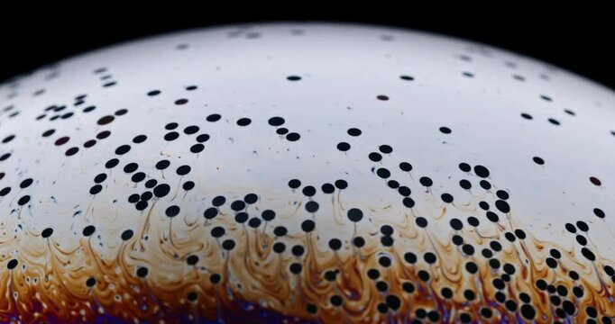countless small dark dots moving around on the complex fluid surface of a soap bubble that is about to burst