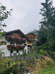 A house with a white roof and a green fence. The house has a balcony with flowers on it. The house is surrounded by trees and bushes