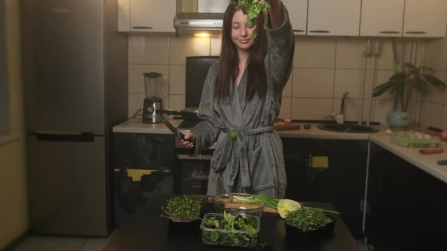 Active young woman in a robe with good mood prepares vegetarian dinner in the kitchen, tossing salad greens into a bowl and dancing joyfully with knife