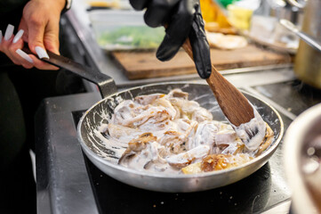 A chef's hand stirs a pan of creamy mushroom sauce using a wooden spoon. The background features kitchen prep items, emphasizing culinary creativity and skill in a professional kitchen setting.
