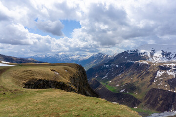 River gorge between high mountains covered with snow. Green grass in front of the cliff. Mountains, rocks, blue sky with clouds in the background