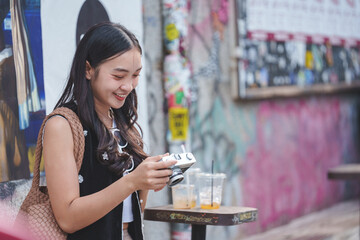 Smiling young asian woman enjoying a refreshing drink while using a vintage camera, surrounded by vibrant graffiti art in a lively urban setting, capturing moments of culture and style