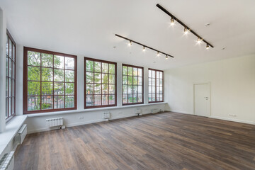 A spacious empty room with panoramic windows in the mansion. White decor, dark laminate. Unfurnished. Courtyard with green trees outside the window.