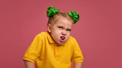 Portrait of emotive preschool girl, child in yellow shirt and braided buns, making funny faces, grimacing, playing against pink studio background. Concept of childhood, emotions, lifestyle