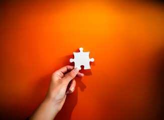 Hand holding a white puzzle piece against an orange background. Symbolizing connection, solution, or missing piece.