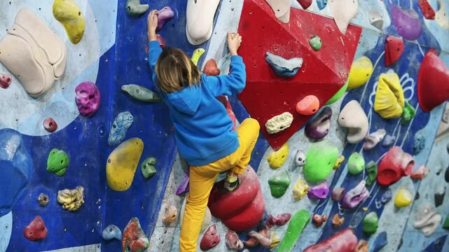 A boy in a blue hoodie climbs an indoor bouldering wall, gripping colorful holds with focus and determination. He reaches for the next hold, showcasing perseverance, strength, and adventure