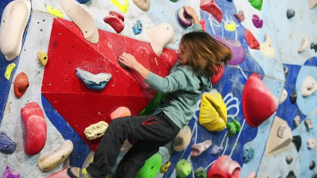 A young boy in a green hoodie climbs an indoor bouldering wall, gripping colorful holds. He focuses on his next move, demonstrating determination, strength, and an adventurous spirit in a climbing gym